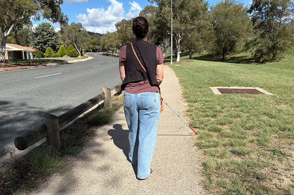 Person walking with a white cane along a suburban footpath, representing Orientation and Mobility assessment and training for people with low vision in Canberra.